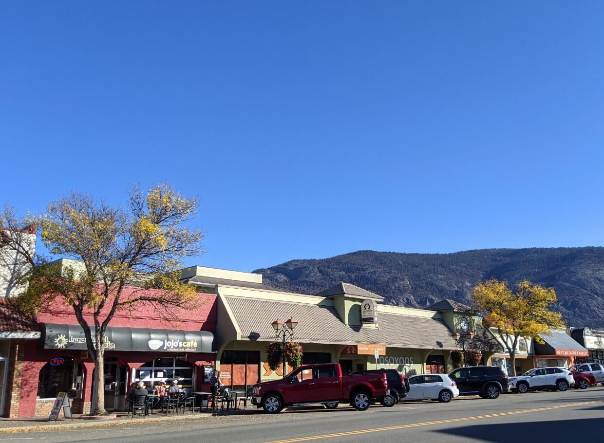 Sidewalk view of downtown Osoyoos with shops, cafes and parked cars on sunny day, with two yellow coloured trees