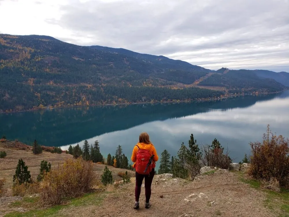Back view of Gemma standing on cliff looking out to Kalamalka Lake with yellow spotted forest on other shore