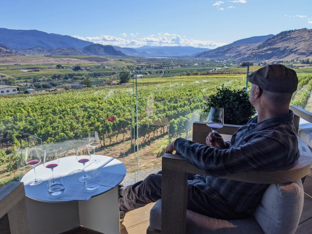 JR sits with back to camera, holding a red wine glass and looks out to scenic view at CheckMate Winery