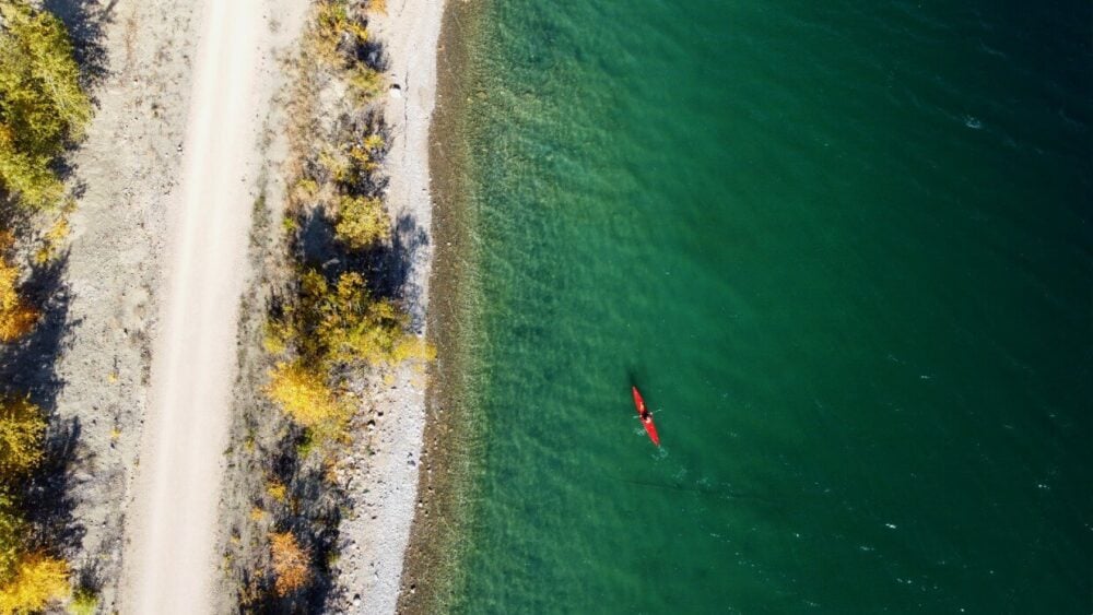 Overhead drone phoo of red kayak on turquoise Kalamalka Lake, next to sandy shore with yellow trees and hiking and biking path