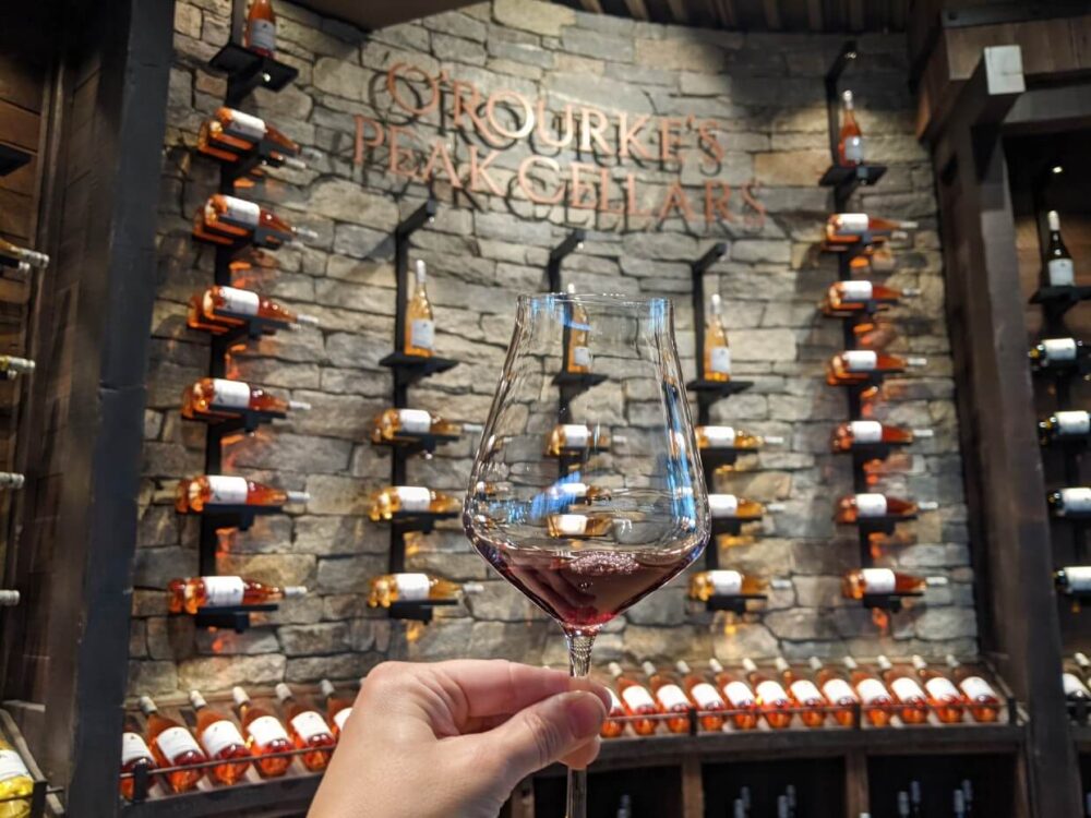 A hand holds up a tasting glass of red wine in front of O’Rourke’s Peak Cellars wine wall filled with horizontal wine bottles