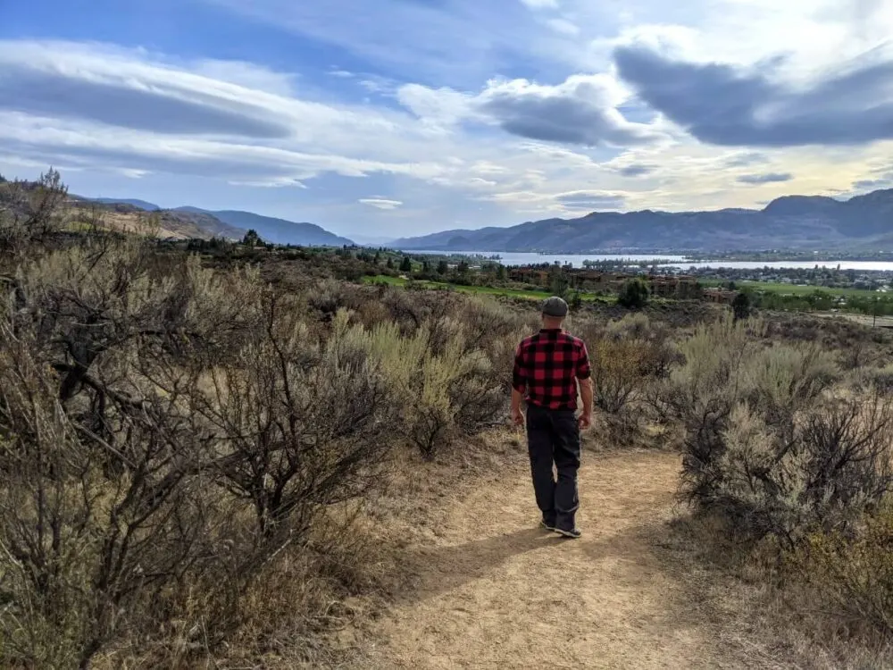 Back view of JR walking away from camera, along dirt path lined with sagebrush, with views of Osoyoos in background