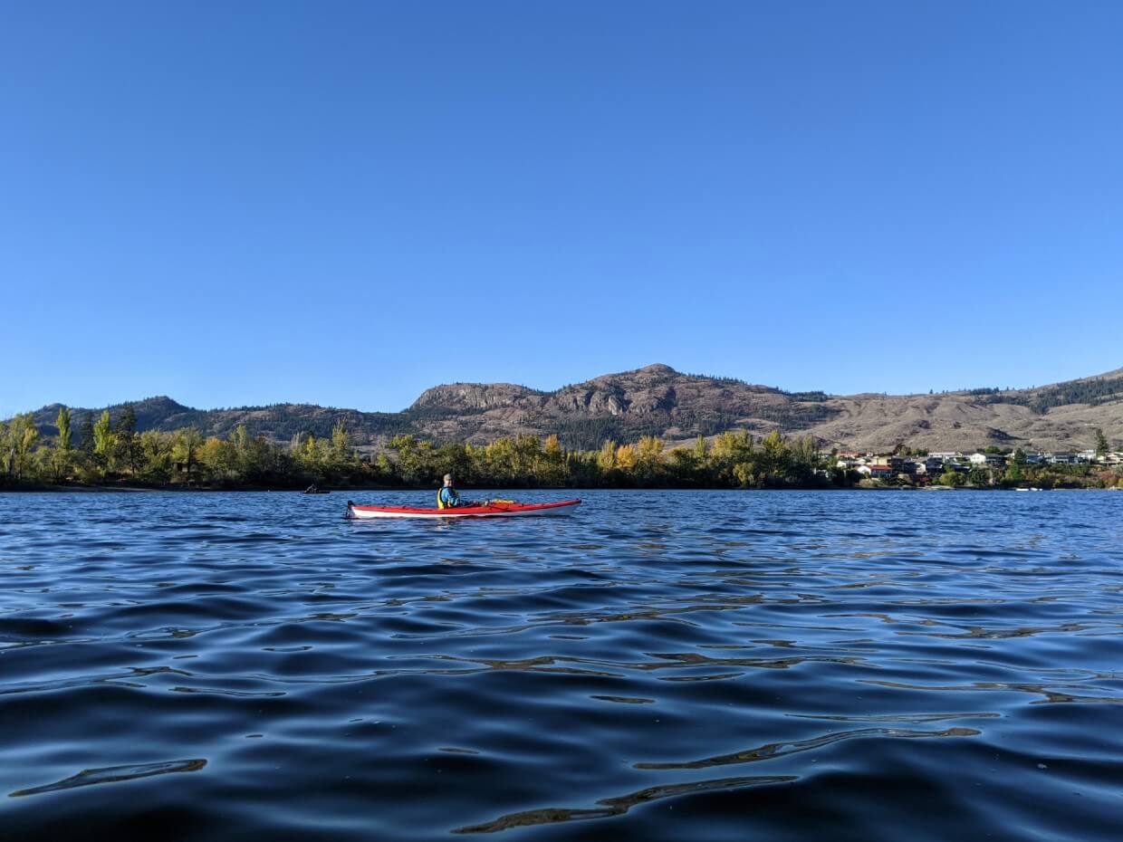 Kayak view of red kayak on Osoyoos Lake, with yellow coloured trees lining shore in background
