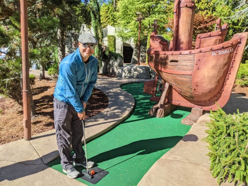 JR looks at camera holding mini golf put, with golfing green behind. There is a large boat as an golfing obstacle behind