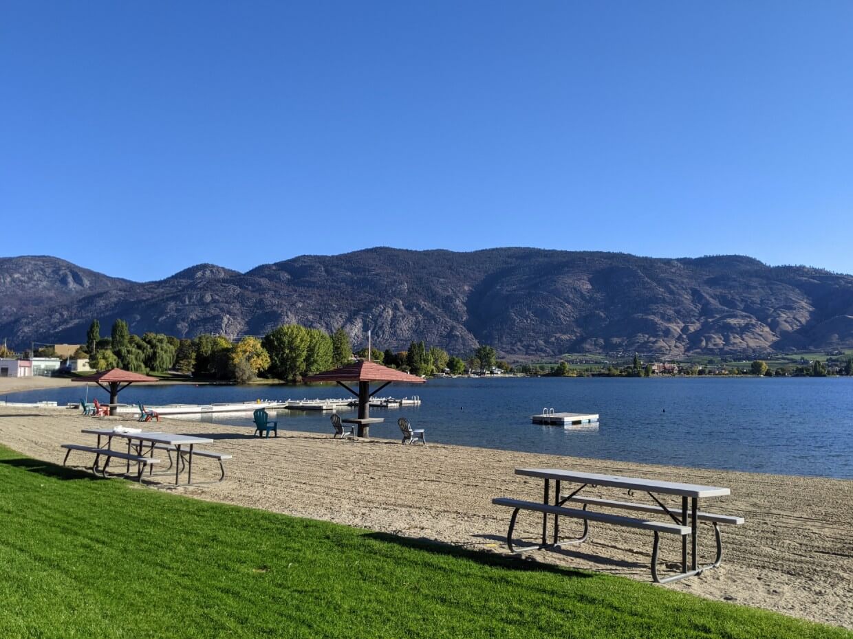 Side view of on beach and lawn space, with picnic tables and Adirondack chairs, next to calm blue lake and surrounding hills