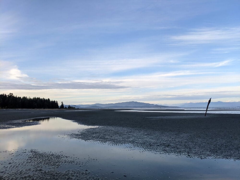 Side view of sandy beach at sunset, with standing water on surface and layers of mountains in background