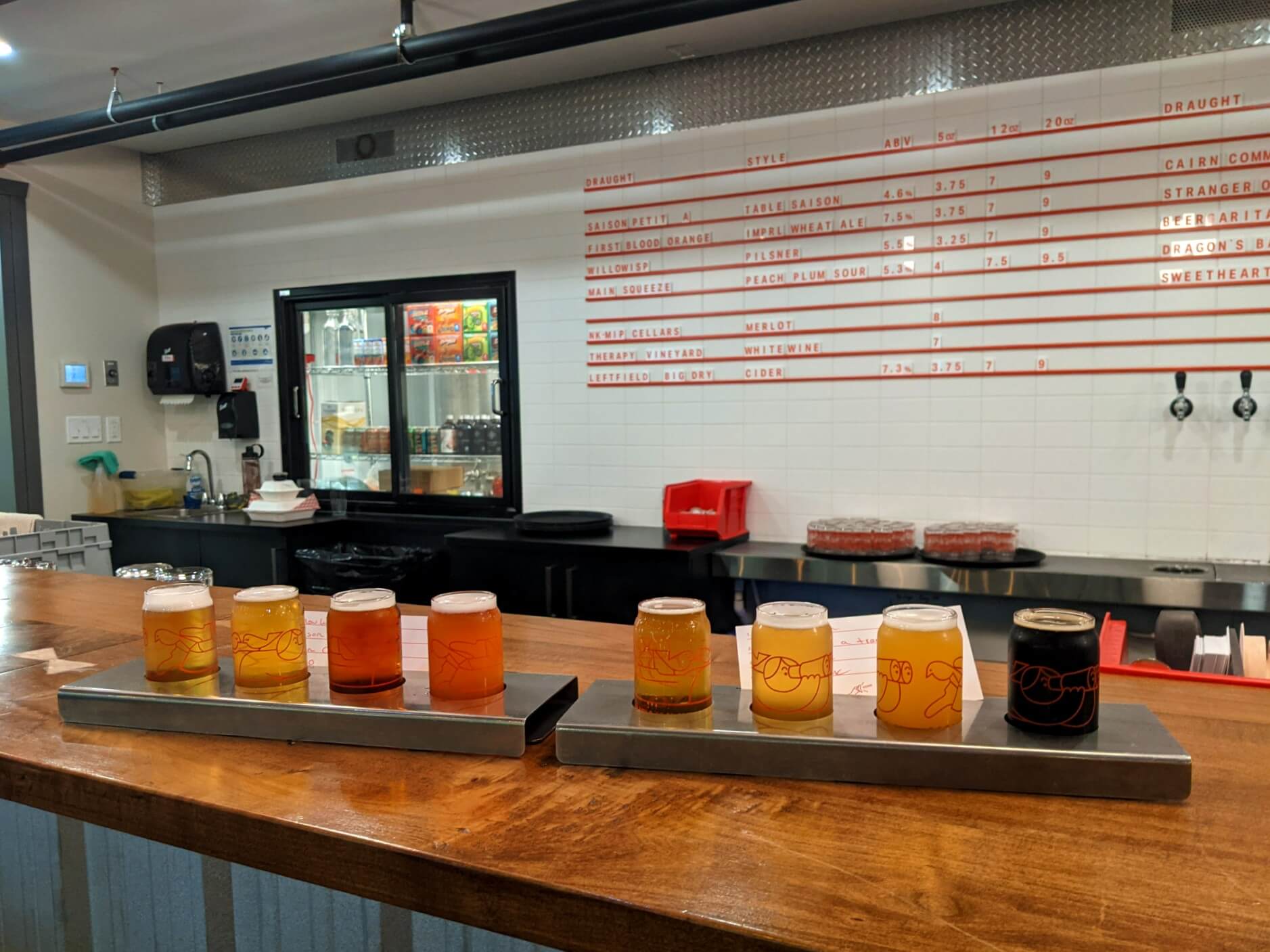Two beer flights with eight different beers total sit on top of the wooden bar at Mountainview Brewery. The wall behind is white and lists the names of the beers