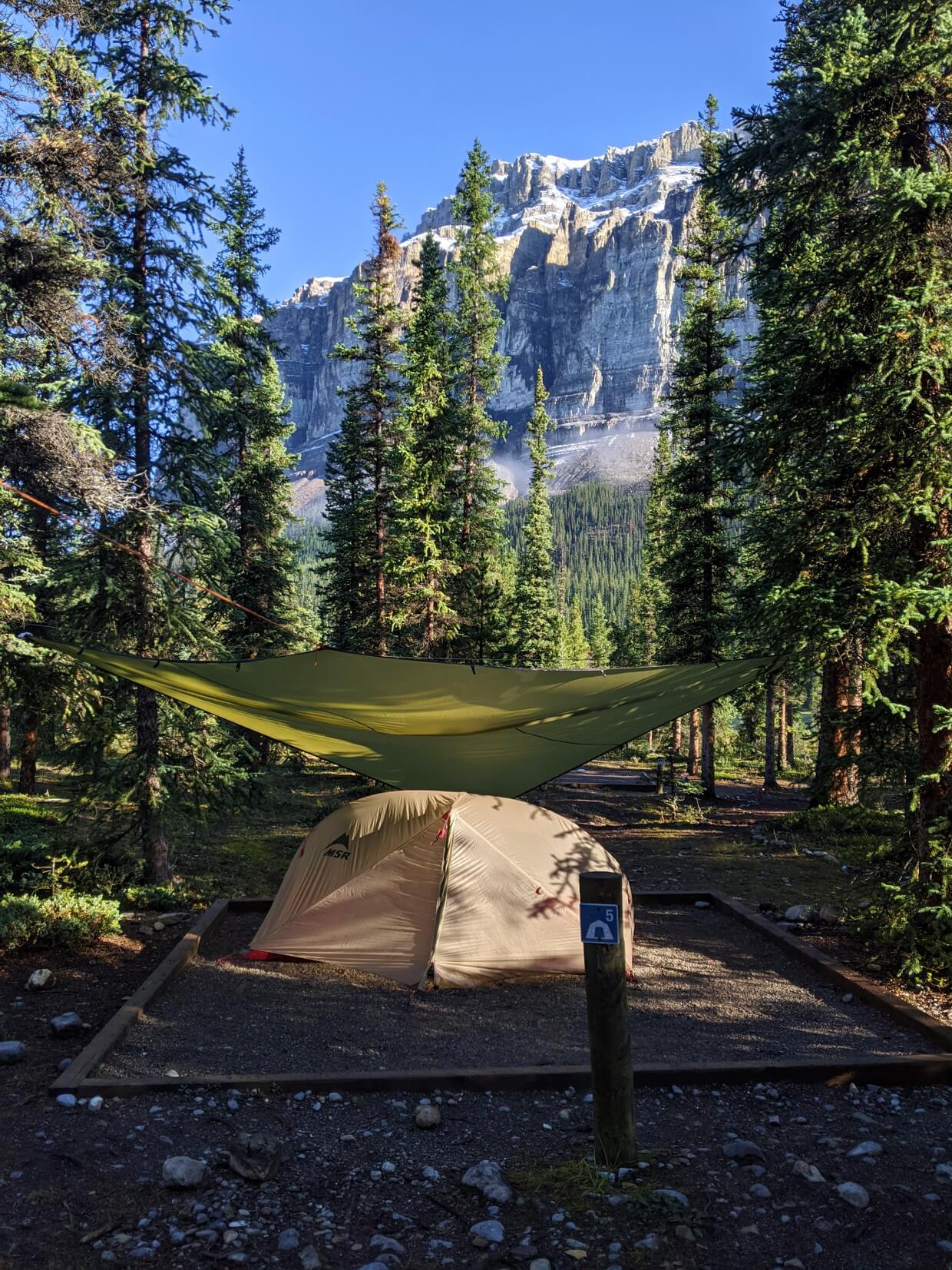 A tent is set up underneath a tarp on tent pad, surrounded by forest with mountains in background