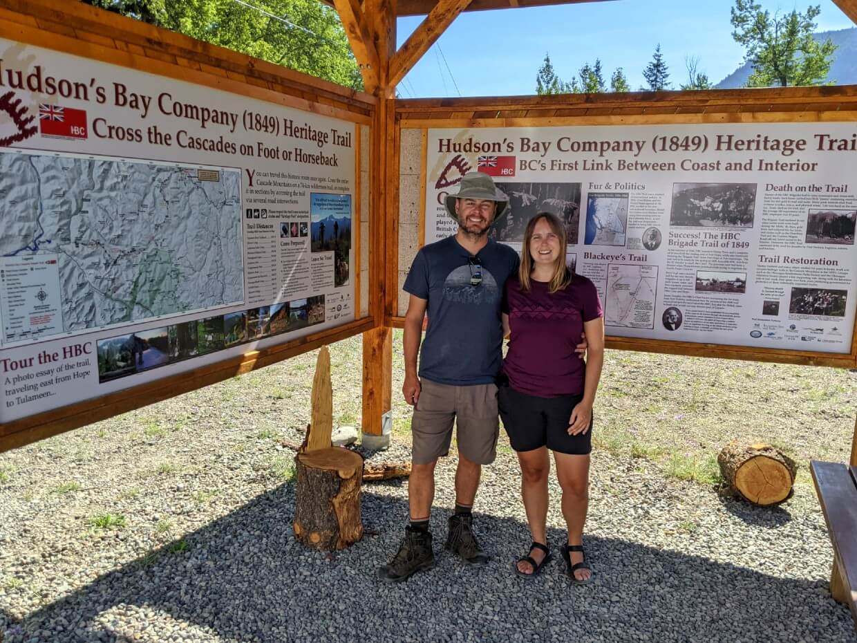 Gemma and JR stand smiling at the camera in front of Hudson Bay Company Heritage Trail signage at the Tulameen Trailhead