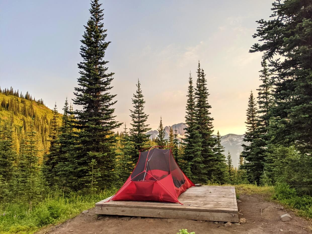 A tent is set up on a wooden tent platform in an open location with some scattered trees and mountain views in the background