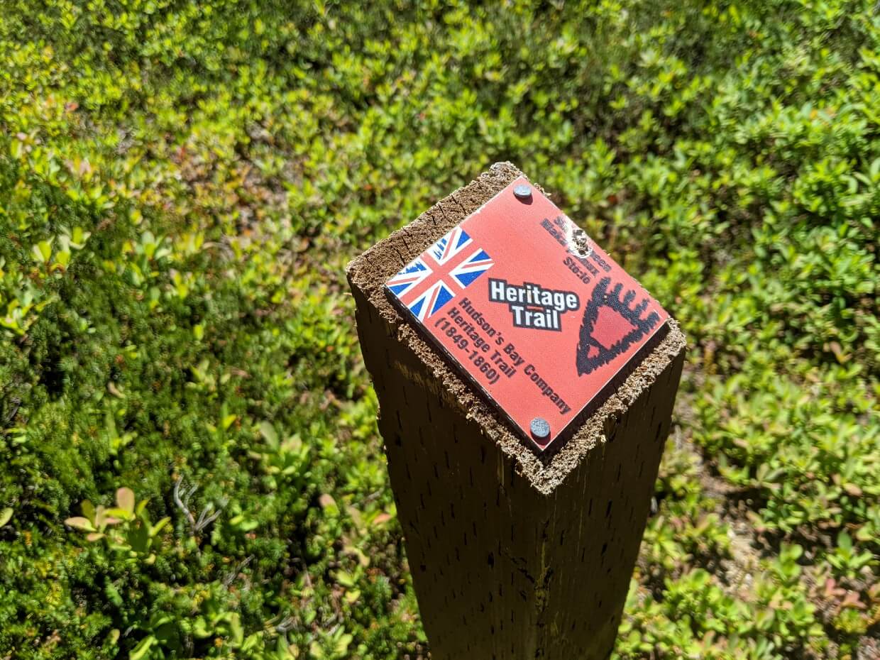 Close up of wooden post on hiking trail, adorned with red Heritage Trail sign, stating Hudson's Bay Company Heritage Trail (1849-1860) with flag and a grizzly bear paw print