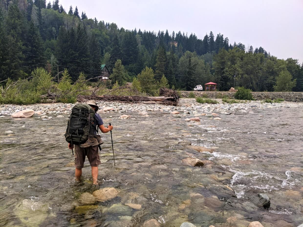 Back view of JR crossing the Tulameen River with hiking pole. Water is half way up his shin. A white van and red roofed kiosk is visible in the background