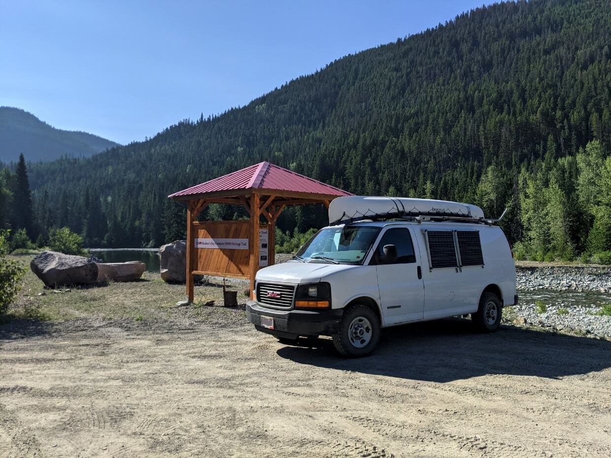 Side view of white van parked at Tulameen Trailhead, with red roofed interpretive kiosk located adjacent