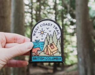 A hand holds up a colourful West Coast Trail patch in front of a forest