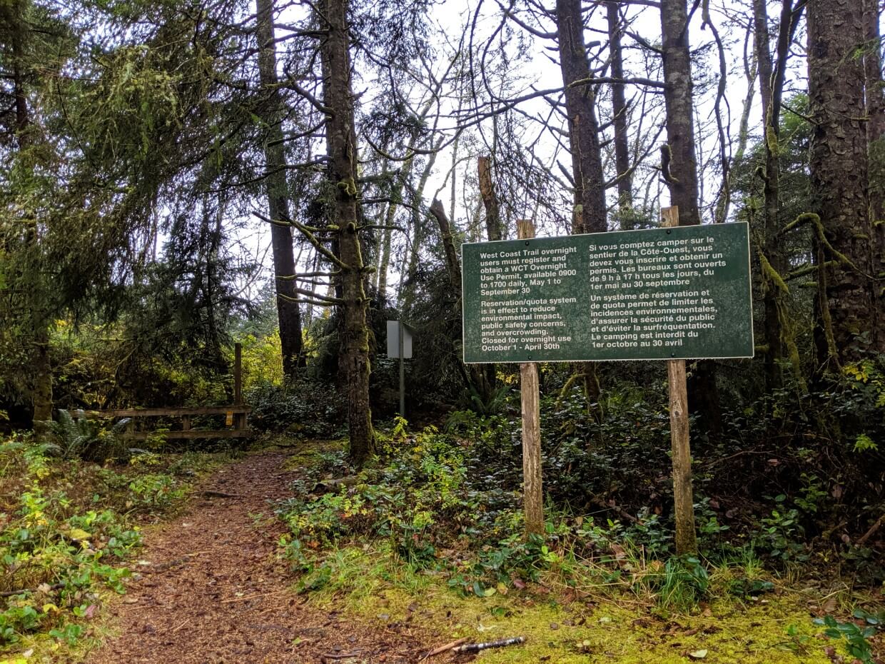 Large green Parks Canada West Coast Trail sign in front of forest and dirt hiking trail