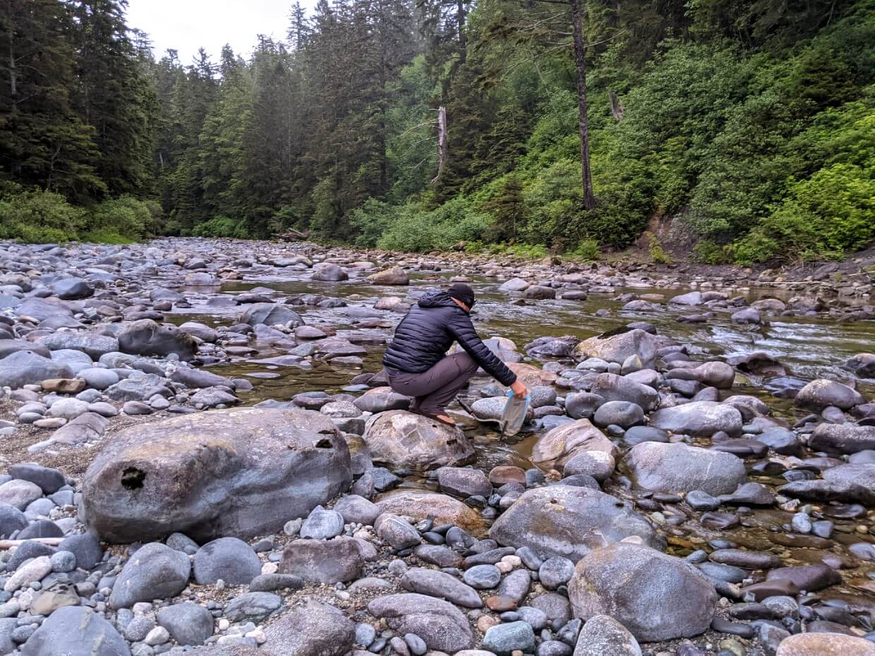 Back view of JR crouching down in creek with hydration bladder, collecting water in Carmanah Creek