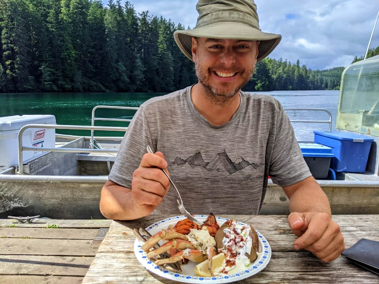 JR facing camera at picnic table, digging into seafood lunch at the Crab Shack. Half of a crab is visible on his plate, with a baked potato. Behind him is a boat and a view of Nitinaht Narrows