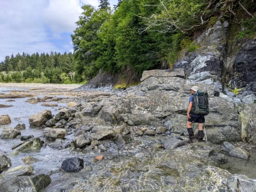 Back view of JR hiking around a rocky headland with the sandy shores of Pachena Bay visible behind