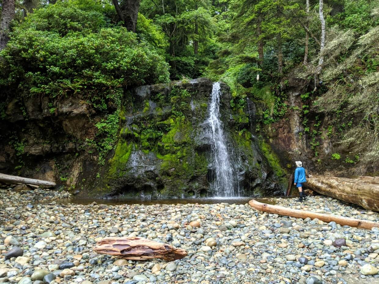 JR stands on rocky beach looking up at Bonilla waterfall, which is cascading from a mossy rock