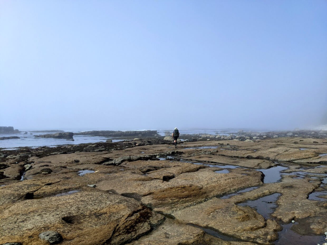 Distant view of JR hiking away from camera on rocky shelves, with ocean in background