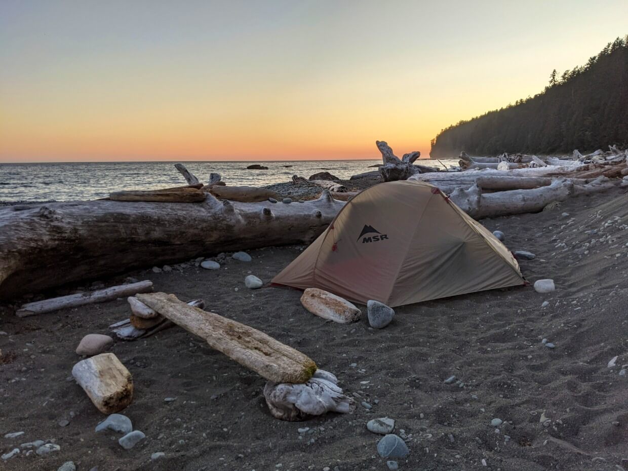Sunset colours in sky behind set up tent on sandy beach, surrounded by driftwood
