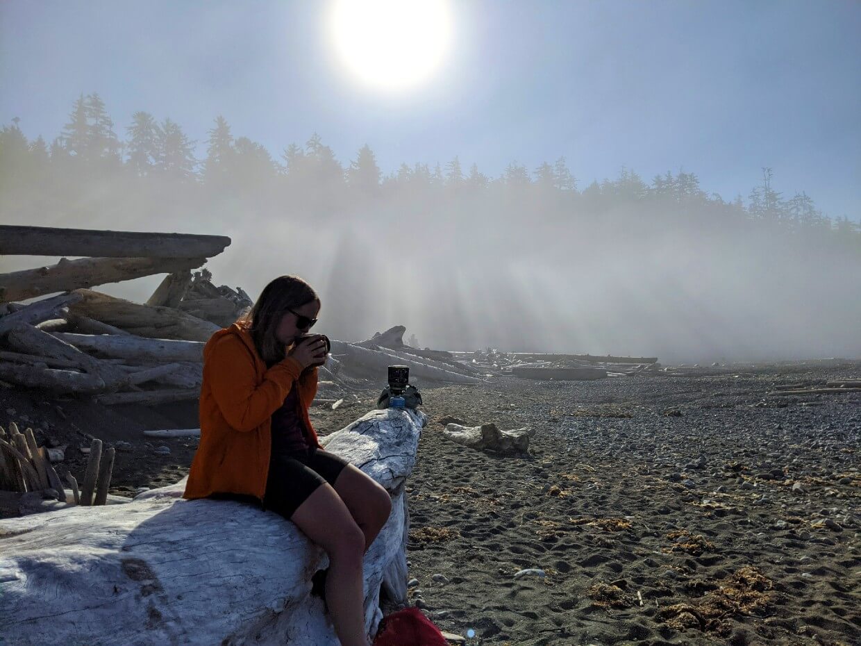 Gemma is sat on driftwood log on sandy beach, with sun streaming through fog in background