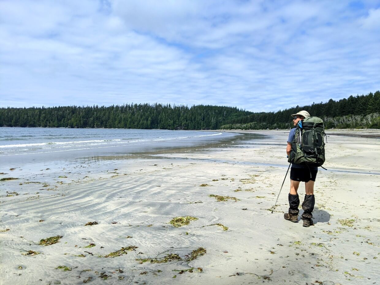 Back view of JR walking down sandy Pachena Bay beach with backpack on 