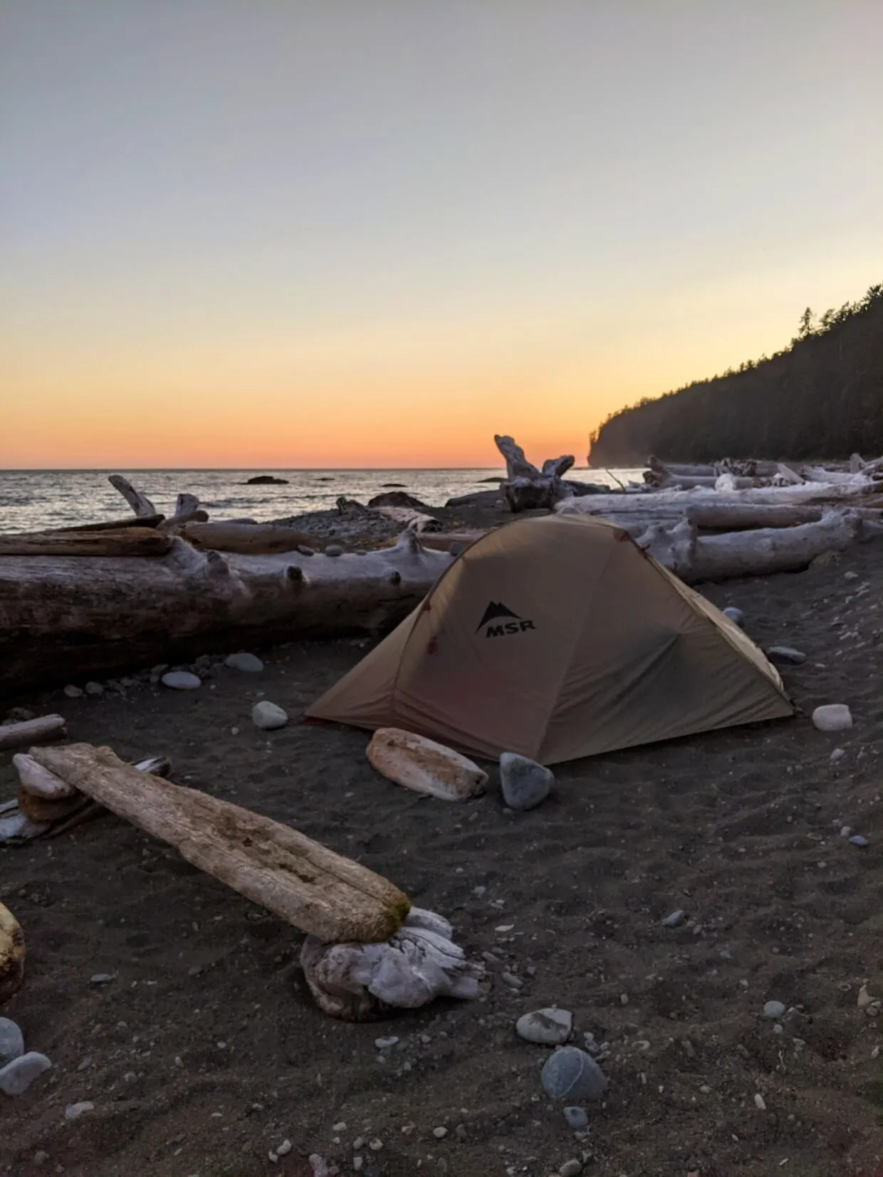 Sunset colours in sky behind set up tent on sandy beach, surrounded by driftwood