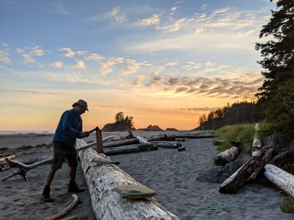 JR standing and chopping wood on large driftwood log during sunset on First Beach, with orange colours in sky