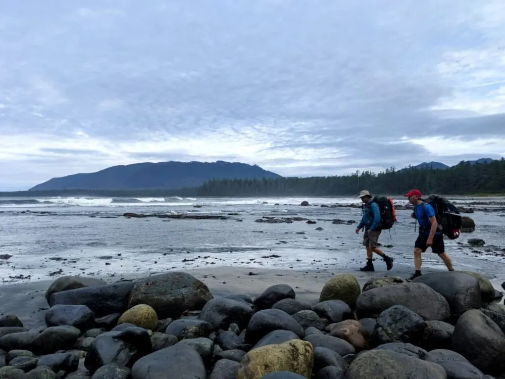 Side view of JR and John hiking beach section of the Nootka Trail, with ocean and mountains behind and rocks in foreground