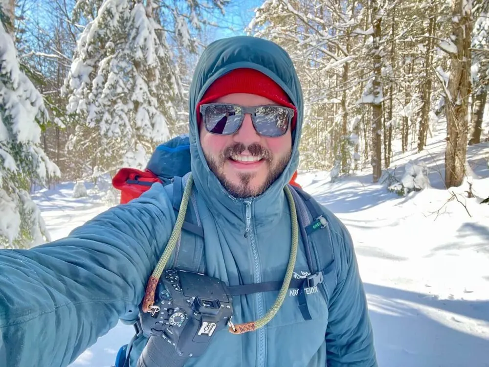 Thomas Coldwell selfie with winter background featuring trees and snowy ground. Thomas is wearing a blue insulated jacket, sunglasses and a red toque