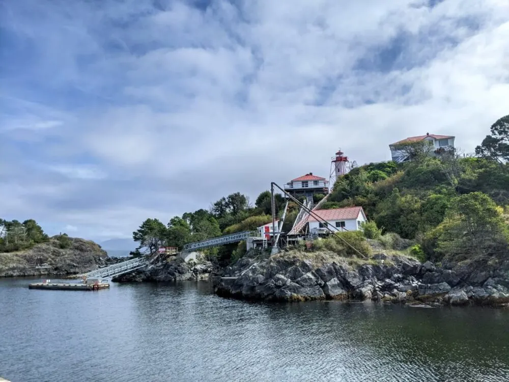 Nootka Lighthouse complex from Yuquot dock, which includes multiple two story buildings and lighthouse perched on rock with dock leading to water