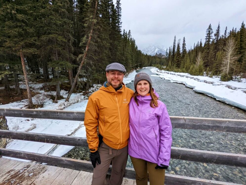 JR and Gemma standing on bridge above partially frozen Spray River, with now capped mountain in background