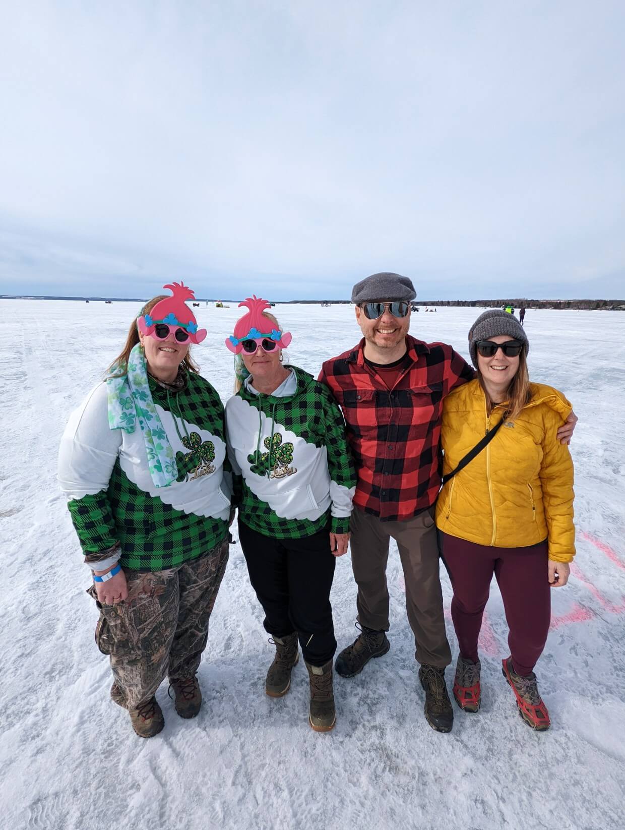 Four people standing on frozen ice looking at camera. Two are wearing matching green checked outfits and themed glasses