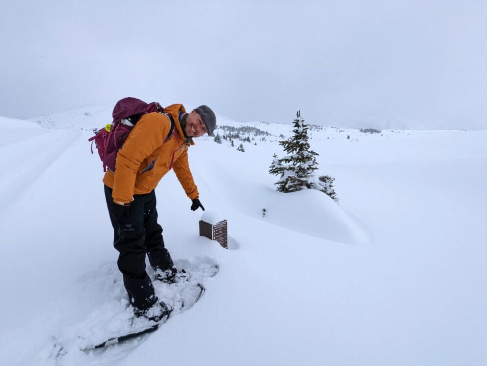 JR is standing and pointing down at a wooden signpost in snowy landscape. The top of a tree is visible