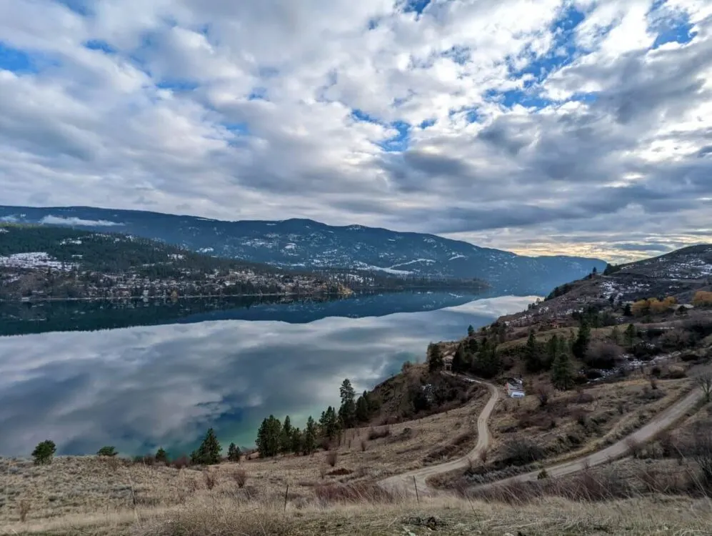 Looking down from elevated viewpoint towards winding road heading towards calm surface on Kalamalka Lake, which is reflecting the surrounding hills and clouds