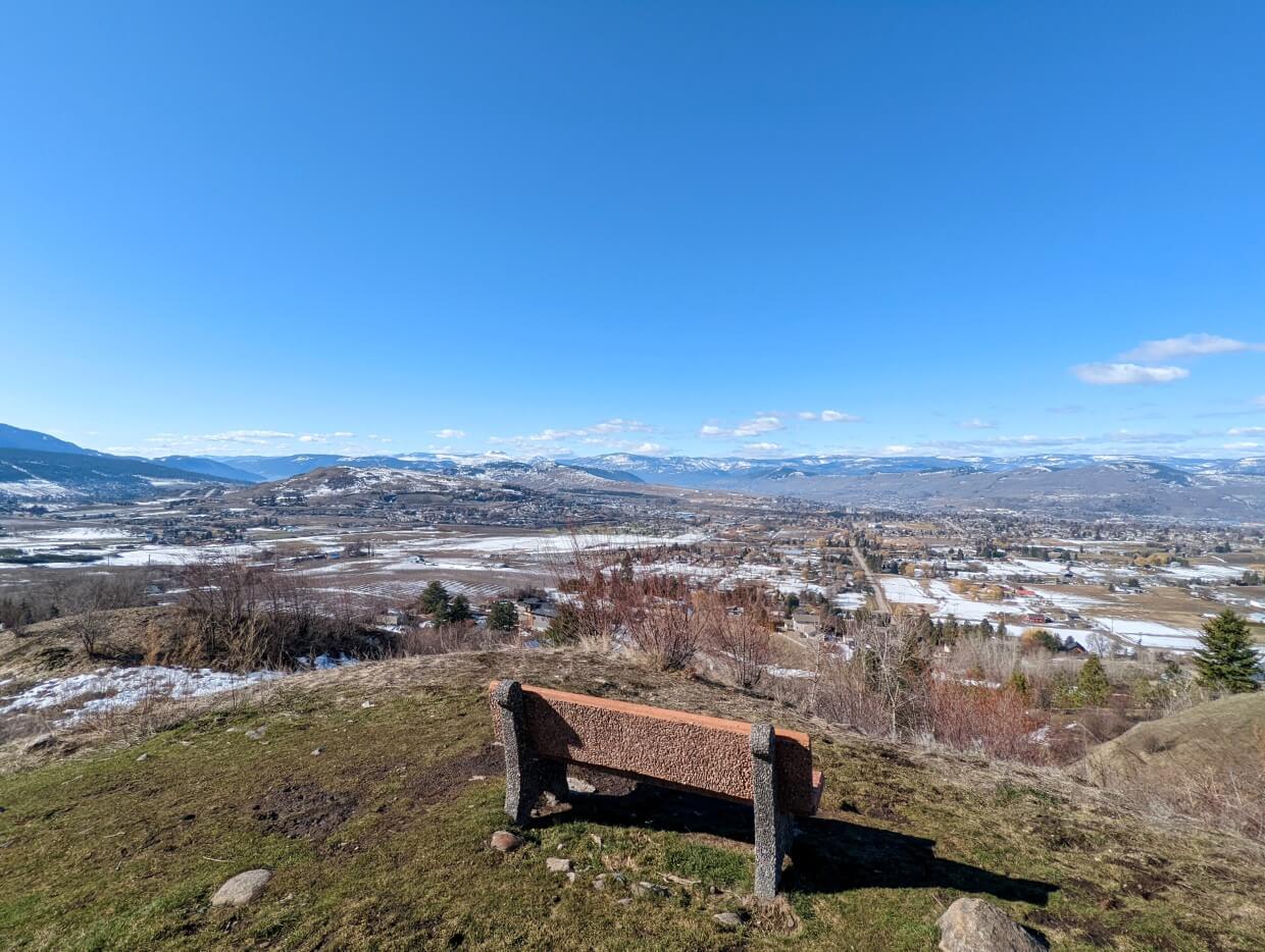 Back view of bench looking out to panoramic view of Vernon from Grey Canal, with snowy fields visible below as well as rolling hills