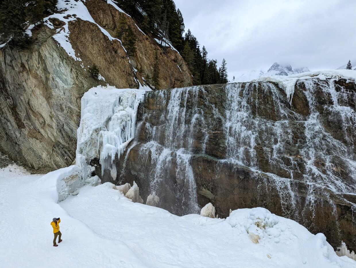 Huge half frozen waterfall cascading over rockface. Gemma stands at the bottom wearing a yellow jacket and taking a photo of the waterfall above