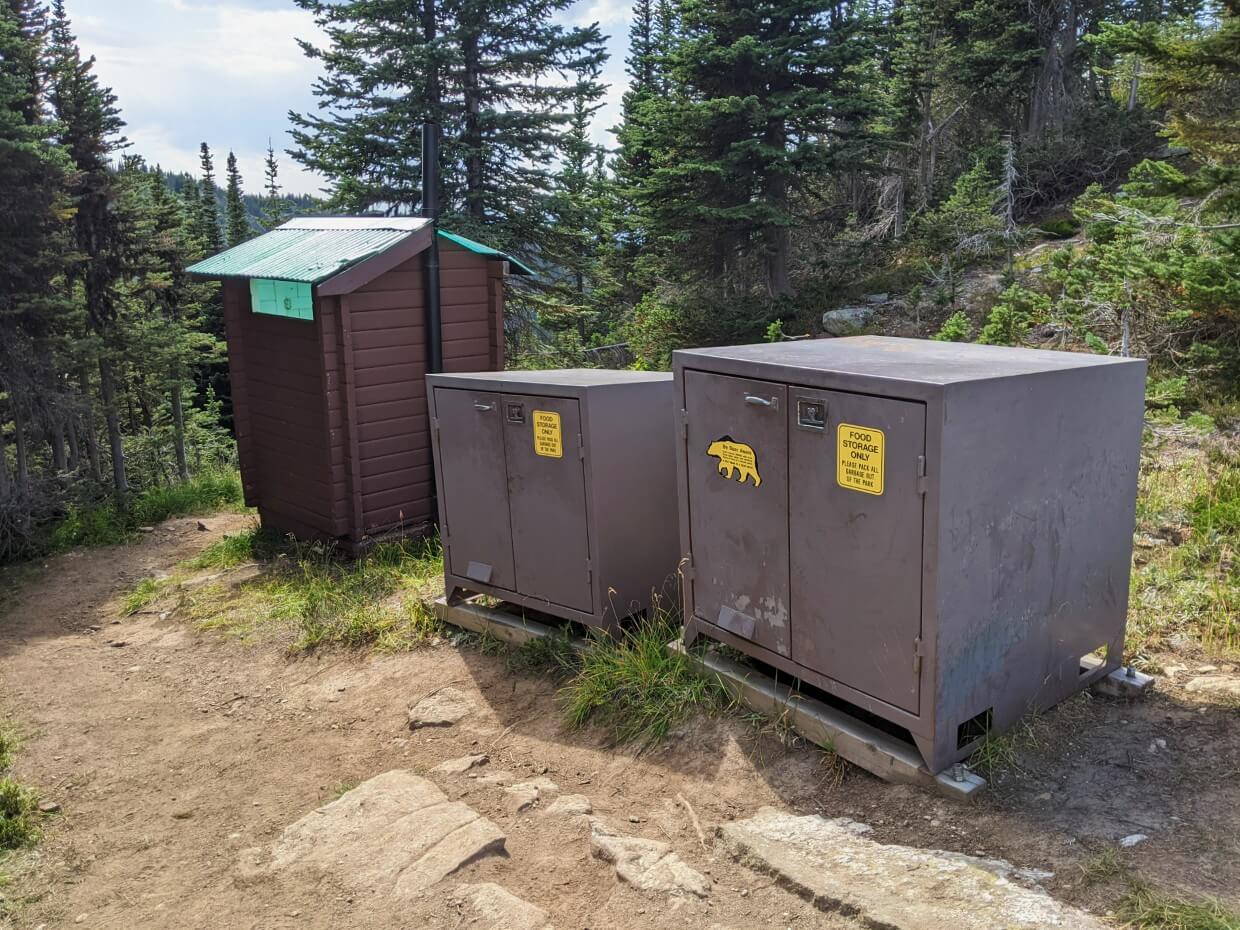 Two large metal bear caches with doors on the front next to wooden outhouse at Sheila Lake campground