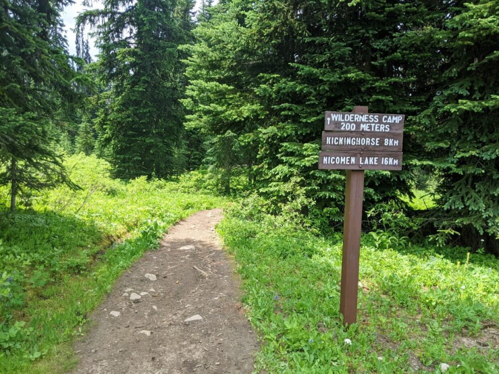 Dirt trail leading into forest, with wooden signage on right - Wilderness Camp 200m, Kicking Horse 8km, Nicomen Lake 16km