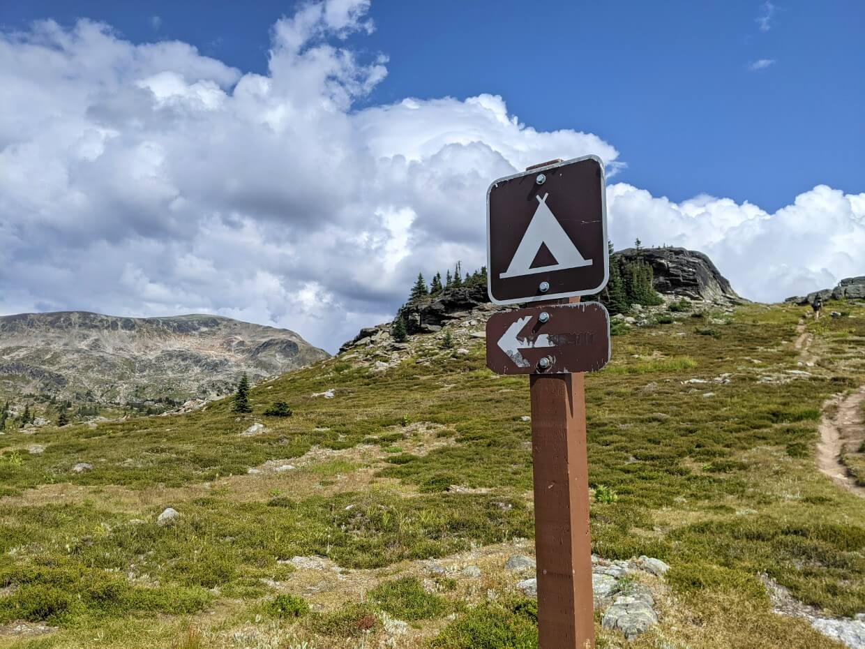 Wooden campground sign with tent symbol and arrow pointing left. Background is sub-alpine scenery with scattered rocks, lichen and a large rock viewpoint