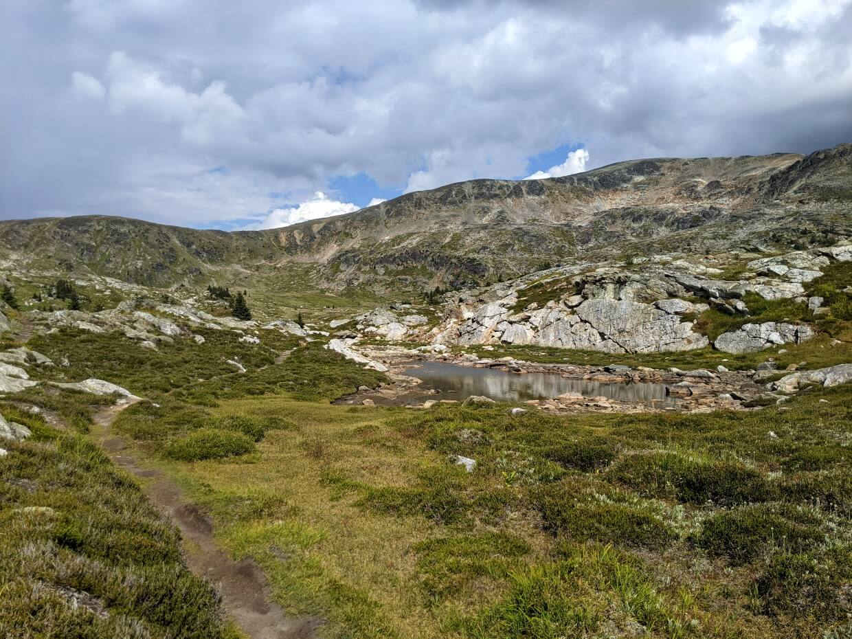 Dirt path leading past small alpine tarn (lake), with background of scattered rocks and mountain ridge