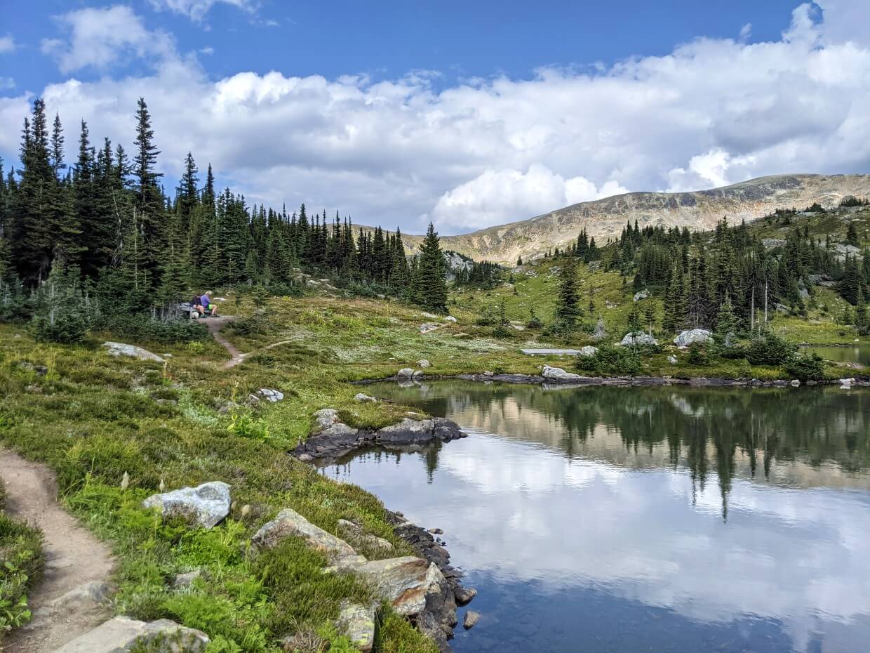 Shoreline of Sheila Lake, with calm reflective water. Two people are sat on a wooden tent pad on left eating lunch. There are scattered trees