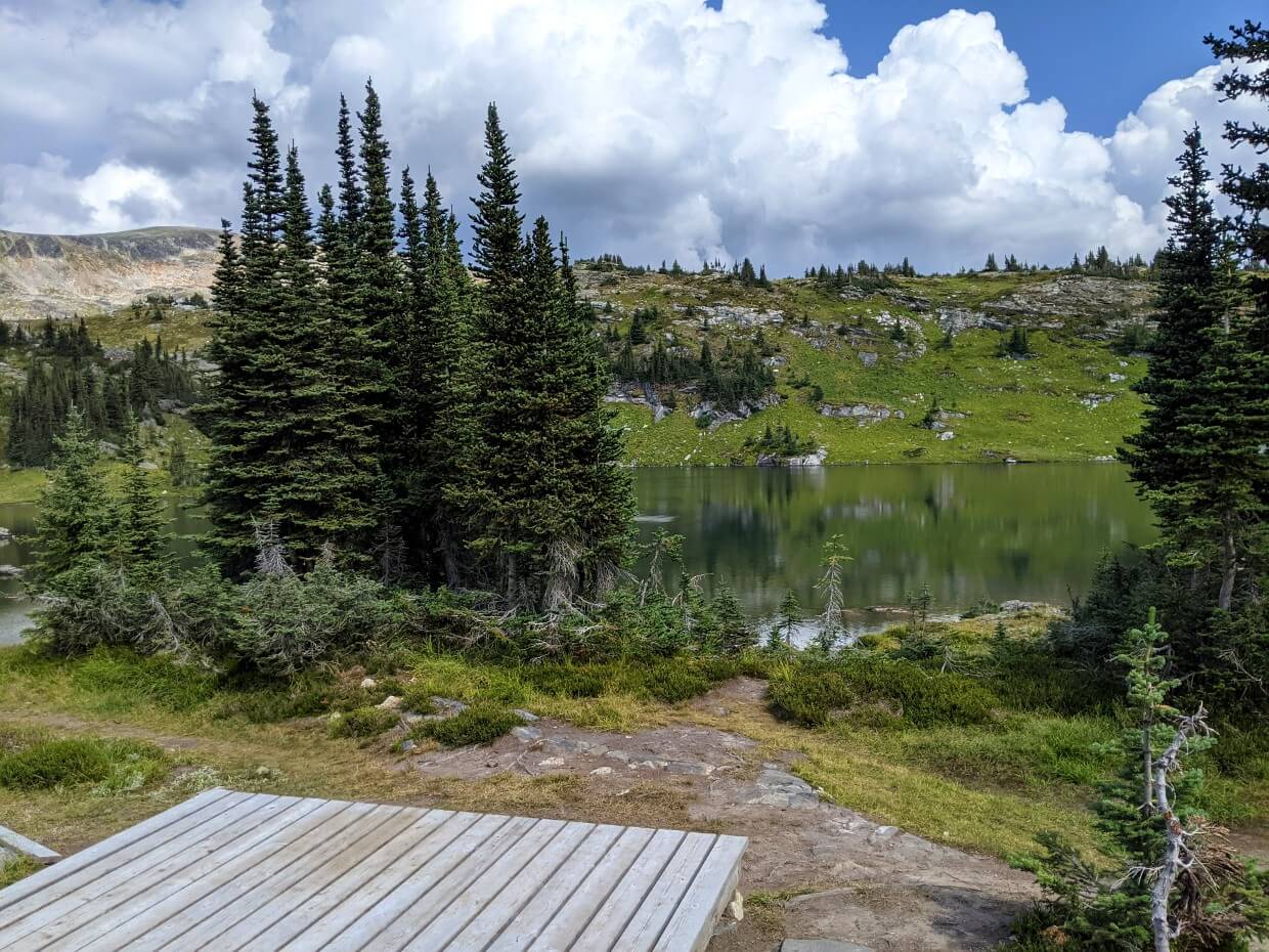 Partial view of wooden tent pad in front of calm Sheila Lake, with scattered trees on right hand side
