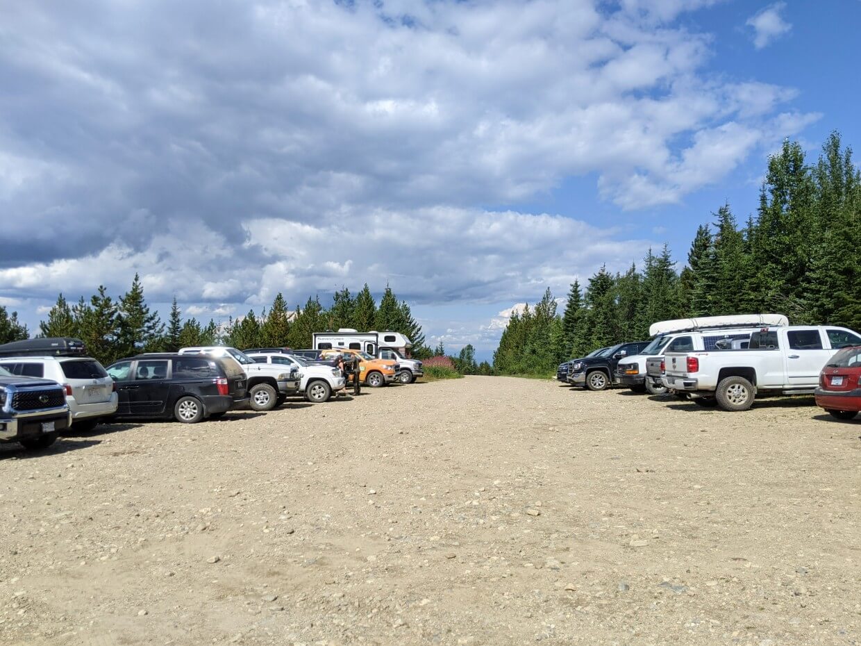 Dirt parking lot with around 18 vehicles parked on left and right. Trees surround the parking lot