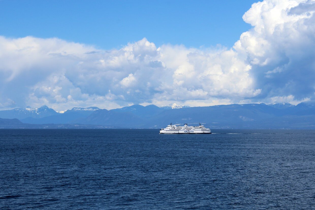 View of a ferry out in the middle of the water. A mountain range lies at the other side of the large body of water. The sky is a mix of clear blue and puffy white clouds.