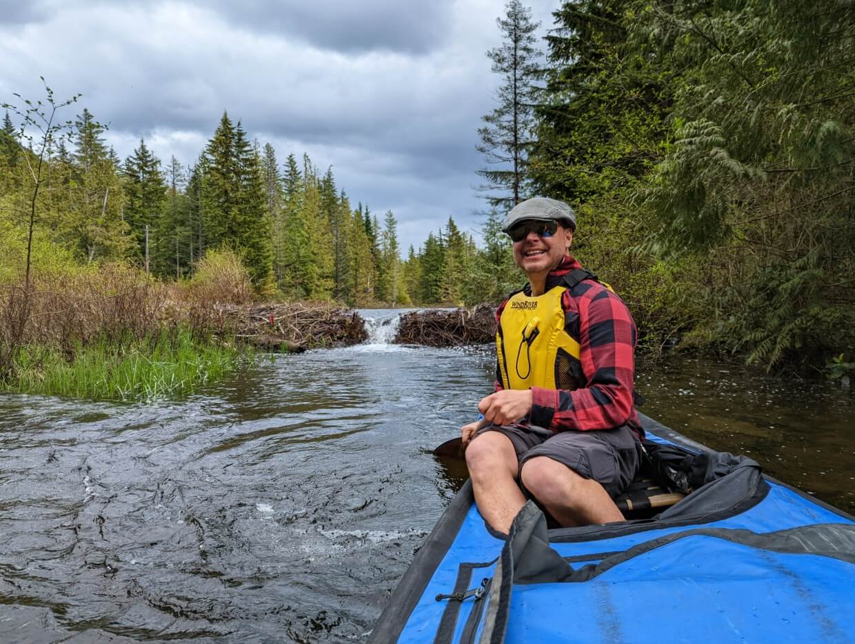 Man smiling at the camera as he paddles in a canoe. Just behind him is a beaver dam on the ponds near Twin Lakes.