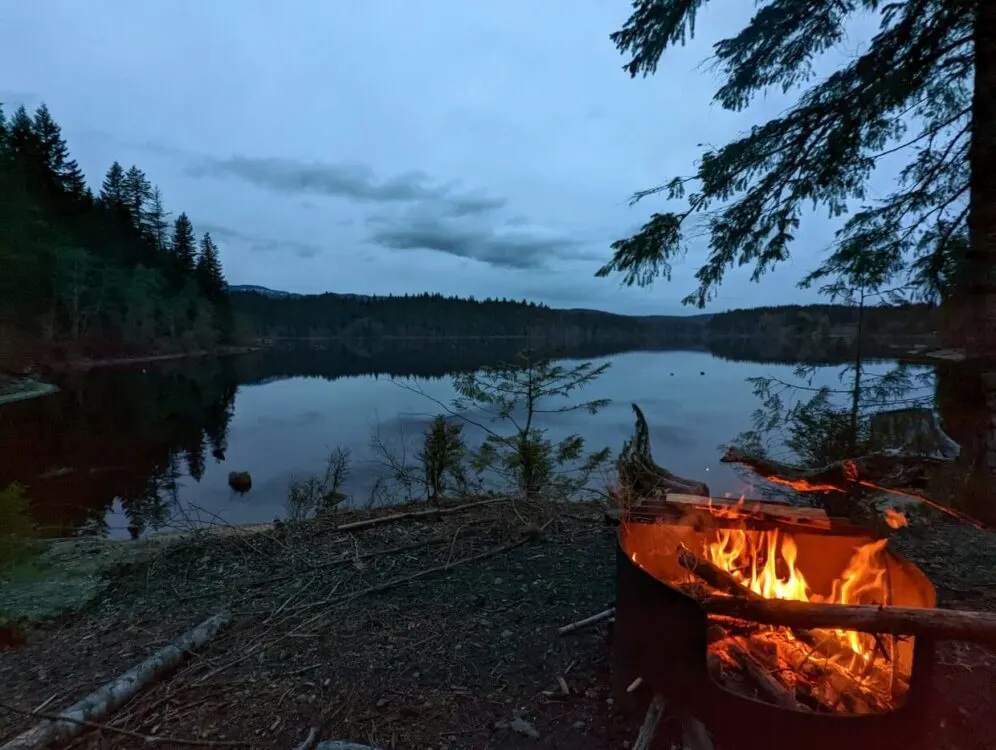 A campfire blazed in a pit near the edge of Fry Lake at dusk. The trees around the lake are silhouetted in the darkening light and are reflecting in the water.