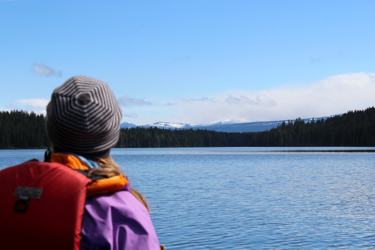 Back of a woman's head who is paddling on a lake. She's looking across the lake at the forest and snow capped mountains.