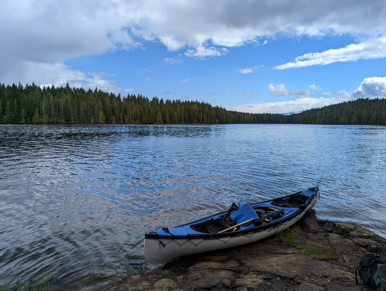 A canoe sits on the edge of a rocky shore on Gosling Lake. The lake is rippling and on the other side of the lake is dense forest.